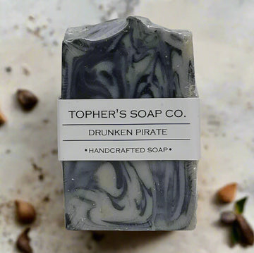 Bar of Tophers Soap Co. Drunken Pirate Handcrafted Soap, featuring dark grey and white swirls, wrapped in a white label with the product name and "Handcrafted Soap," photographed on a light brown speckled surface with scattered botanicals, emphasizing a locally made (Brantford, Ontario) soap with a bold, clove-free Bay Rum aroma.
