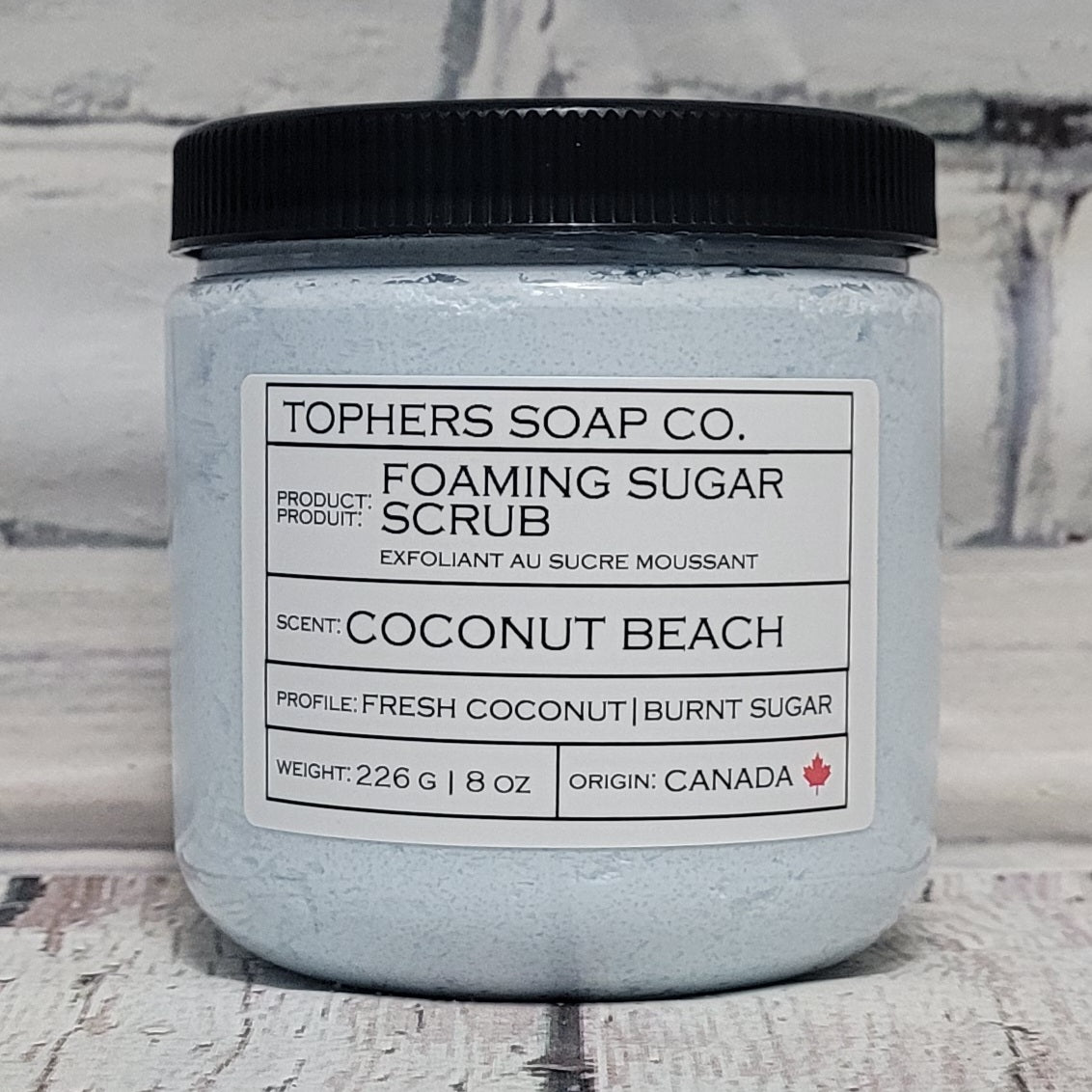 Light Blue sugar scrub in a clear jar with a black lid against a white brick background