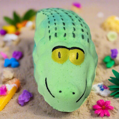 A light green, hand-painted bath bomb shaped like an alligator with black details and yellow eyes, likely scented with Cream Soda, from Tophers Soap Co. in Brantford, Ontario, resting on a sandy surface with tropical decorations.