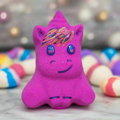 A vibrant pink, hand-painted bath bomb shaped like a chubby unicorn with a colorful mane, glittery eyes, and a smiling face, surrounded by other pastel bath bombs, handcrafted by Tania at Tophers Soap Co. in Brantford, Ontario, Canada.