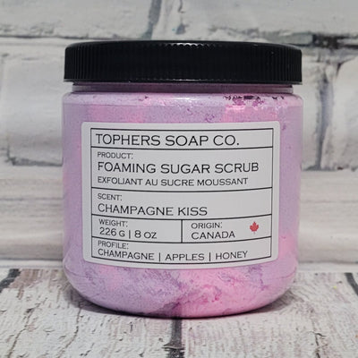 Pink and Purple sugar scrub in a clear jar with a black lid against a white brick background
