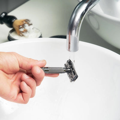 A hand rinsing a white chrome Rockwell 6C Adjustable Double Edge Safety Razor under a bathroom faucet, likely available at Tophers Soap Co. in Brantford, Ontario.