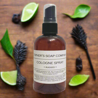 Dark bottle of Tophers Beard Co. Rugged Cologne Spray with a clear spray nozzle and a white label detailing its bold scent, surrounded by lime slices, tobacco leaves, and dark pods on a wooden surface, emphasizing its unique blend and Canadian origin in Brantford, Ontario.