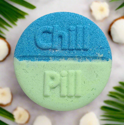 Large, round bath bomb from Tophers Soap Co. in Brantford, Ontario, divided into a textured blue top half with "Chill" embossed and a textured light green bottom half with "Pill" embossed, surrounded by coconut pieces and tropical leaves, showcasing a fun and locally handcrafted bath escape.