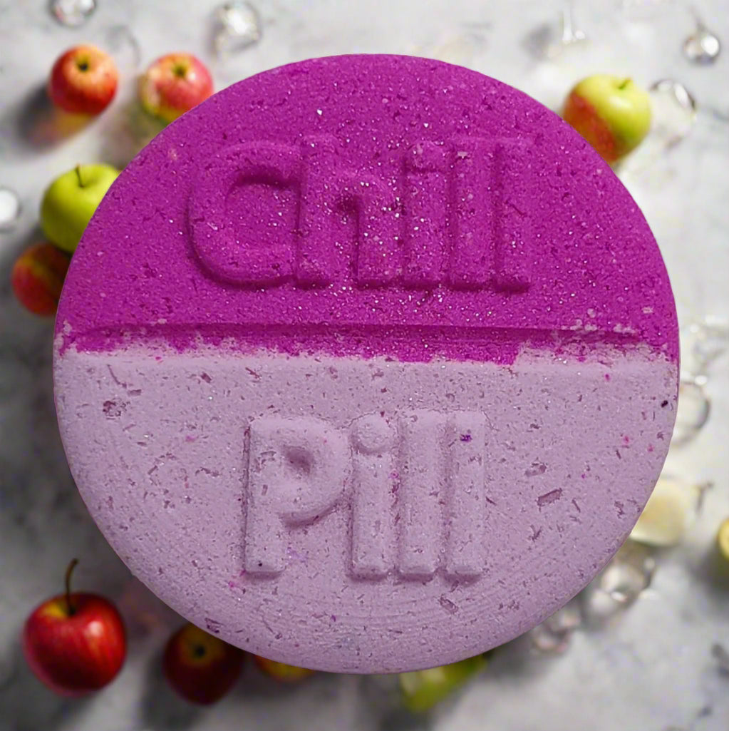 Tophers Soap Co. Champagne Kiss Chill Pill Bath Bomb, a large round bath bomb split in vibrant pink and light purple with "Chill Pill" embossed on it, surrounded by apples and ice cubes, evoking a sparkling champagne, apple, and berry scent, handcrafted in Brantford, Ontario, Canada.