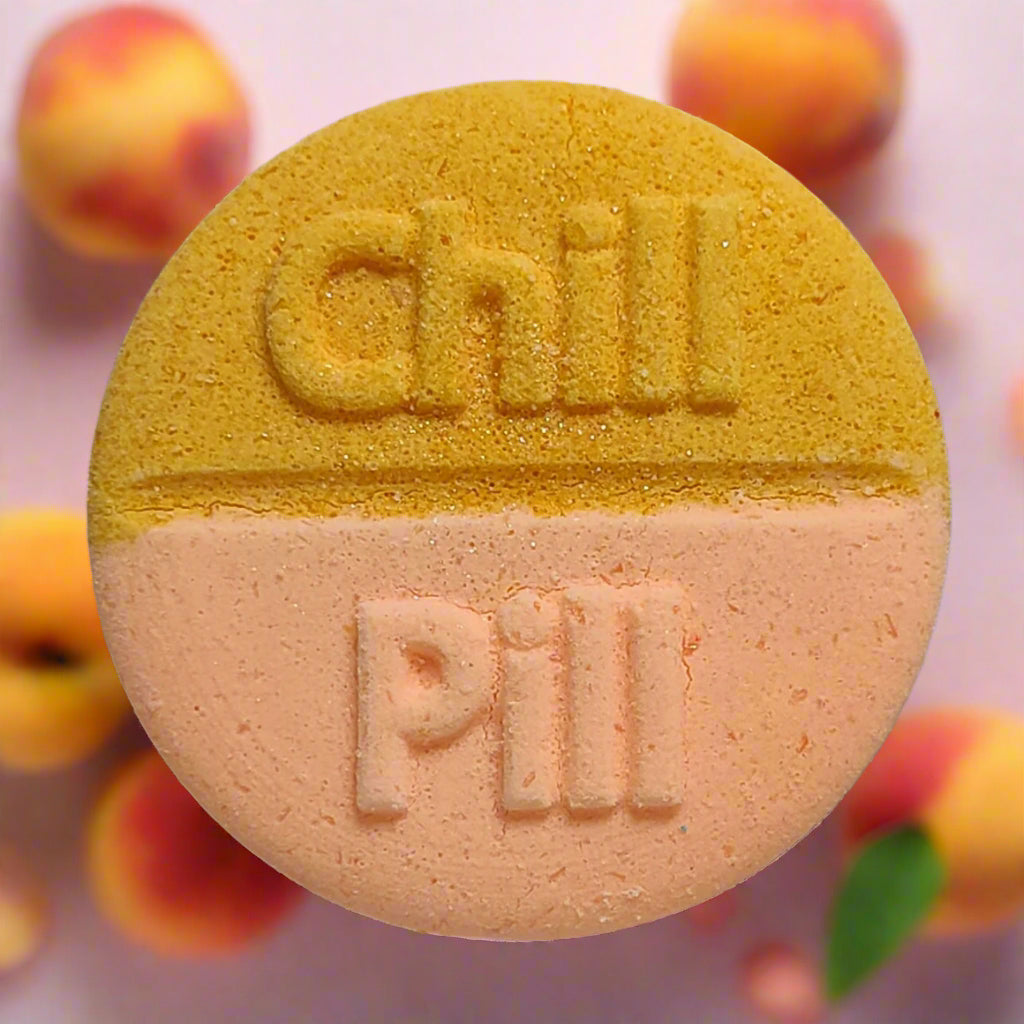 Tophers Soap Co. Happy as a Peach Chill Pill Bath Bomb, a large round bath bomb split in golden yellow and soft peach with "Chill Pill" embossed on it, surrounded by fresh peaches, evoking a sweet and juicy peach scent, handcrafted in Brantford, Ontario, Canada.