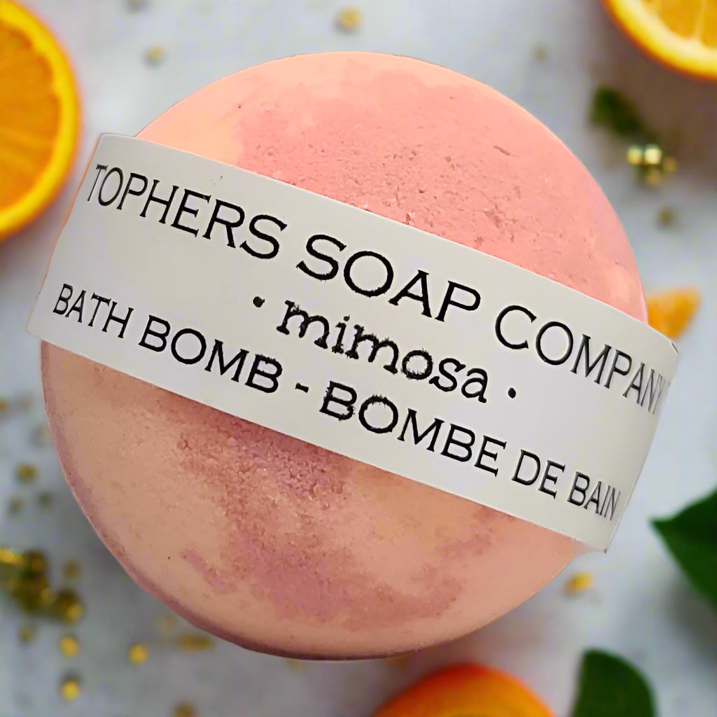 Topher's Soap Company Mimosa Bath Bomb - A swirl of light orange and pink bath fizzer with a label clearly showing the "mimosa" scent, surrounded by orange slices. Made in Brantford, Ontario, Canada.